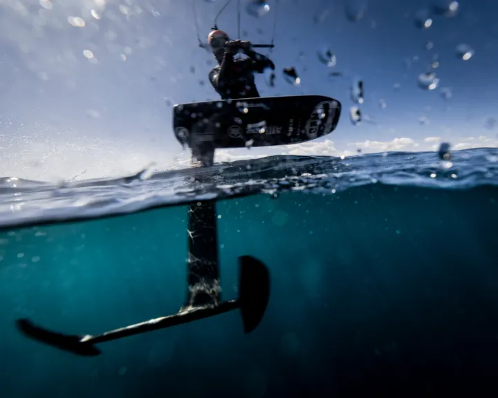 Split-level photograph taken at the waterline. Above it, the underside of the board and a rider against bright sky; below it, the carbon mast and the two wings of the hydrofoil hanging in clear green water.