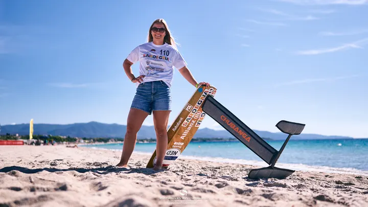 Jessie Kampman standing barefoot on the beach at Quartu Sant'Elena in an event shirt, one hand resting on her board, which is stood on end in the sand with the hydrofoil in the air beside her.