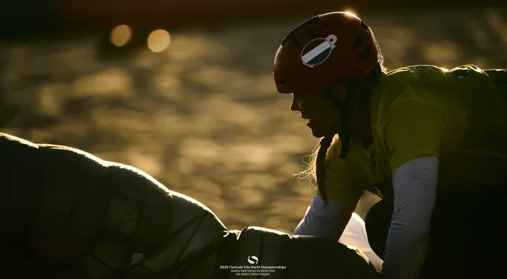 Late afternoon light. Jessie Kampman in a yellow leader's bib and helmet, seen past the out-of-focus leading edge of her kite, everything reduced to gold and shadow.