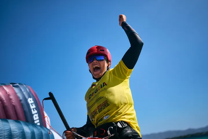 Jessie Kampman with one fist raised and her mouth open, still on the water, the kite's edge crossing the bottom of the frame against a hard blue sky.