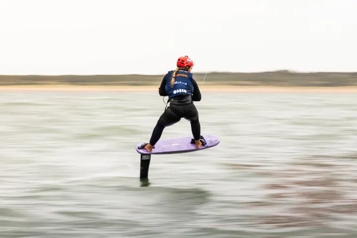 Jessie Kampman riding away from the camera at Brouwersdam, the board lifted clear of grey-green water that has smeared to horizontal streaks under a blank white sky.