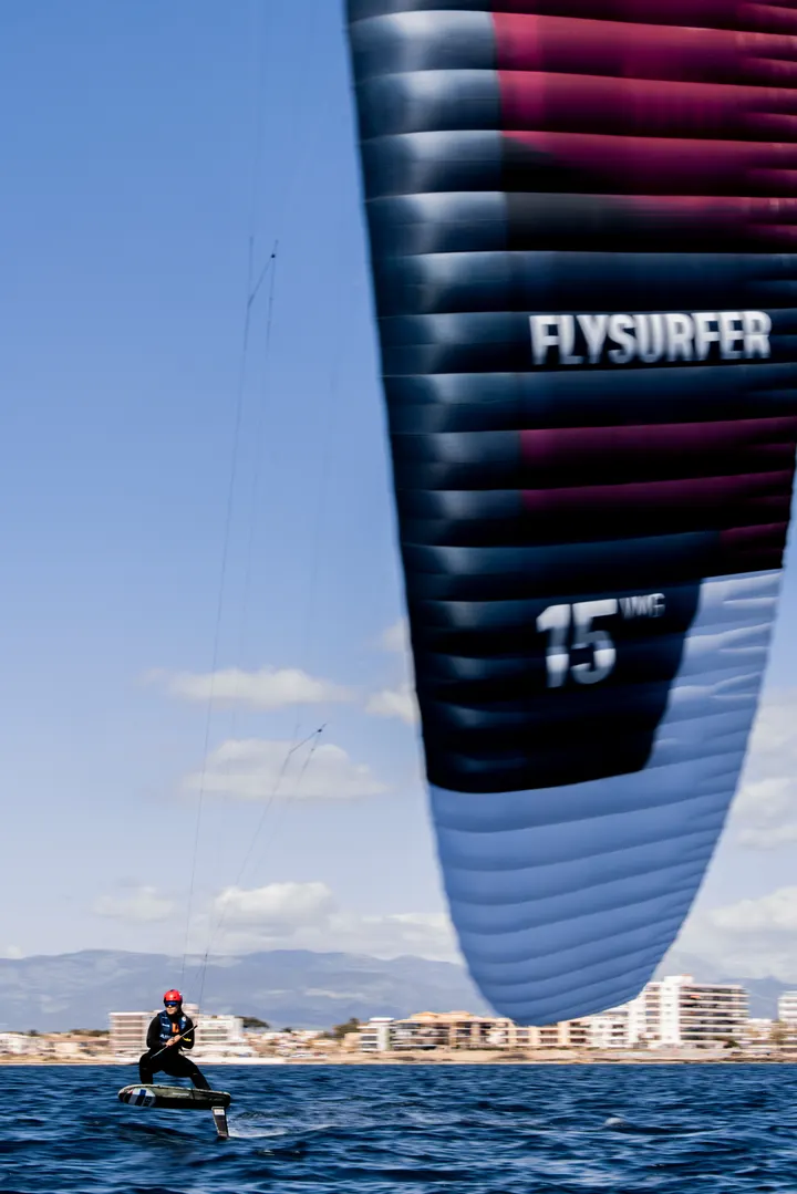 A large kite filling the top of the frame, its trailing edge marked 15 VMG, the rider a small dark shape on the water far below it.