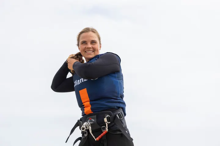 Jessie Kampman on the beach in a wetsuit pulled to the waist, both hands behind her head, tying her hair back, laughing.