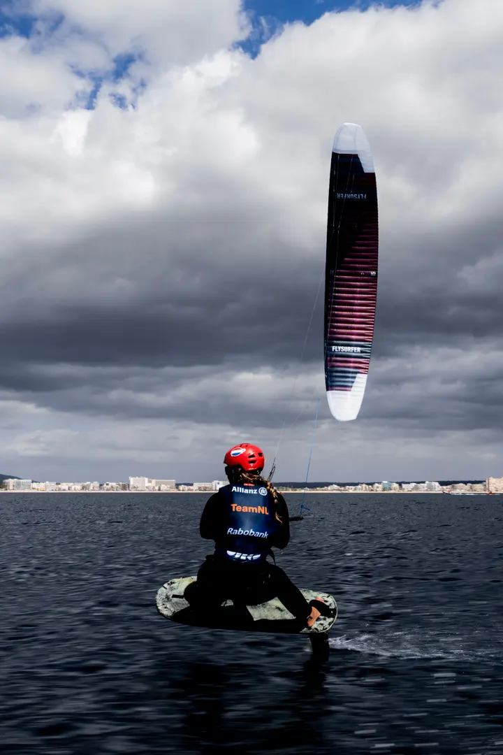 Jessie Kampman sitting in the water on her board under a bank of storm cloud, her kite parked directly overhead, the city skyline on the horizon behind her.