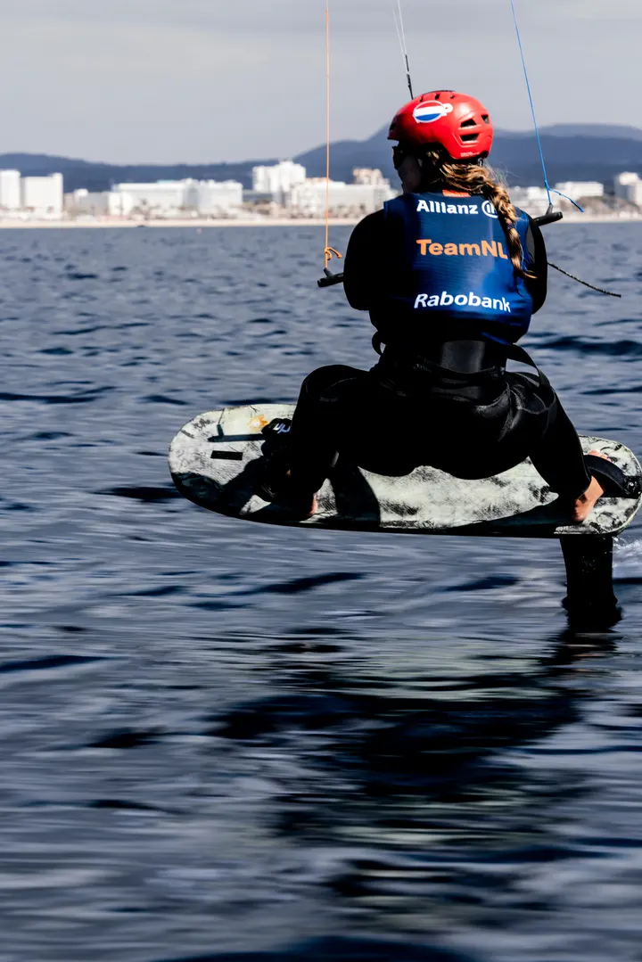 Jessie Kampman sitting on her board in flat water with her legs over the side and the foil below her, stopped, waiting to get going again.