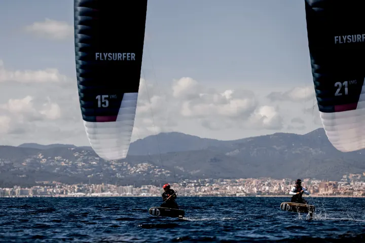 Two riders on the water in Palma under two kites of different sizes, one marked 15 and the other 21, with the city and mountains behind them.