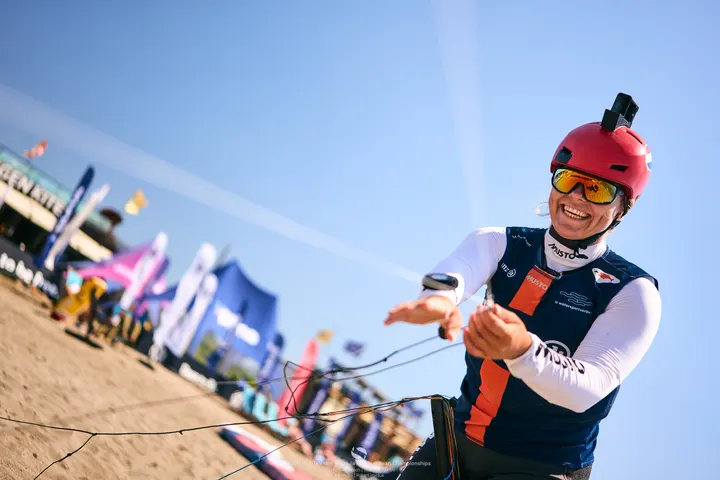 Jessie Kampman laughing on the beach in her helmet and sunglasses, her kite bar in both hands and the lines running off out of frame.